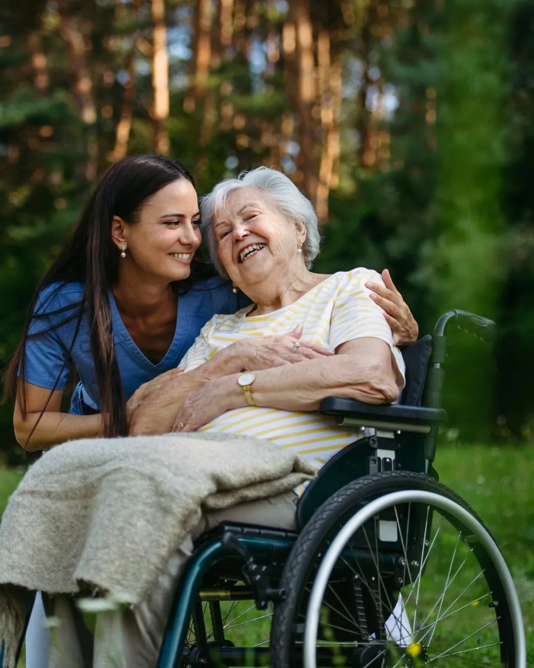 A caregiver hugging a smiling older woman in a wheelchair outdoors