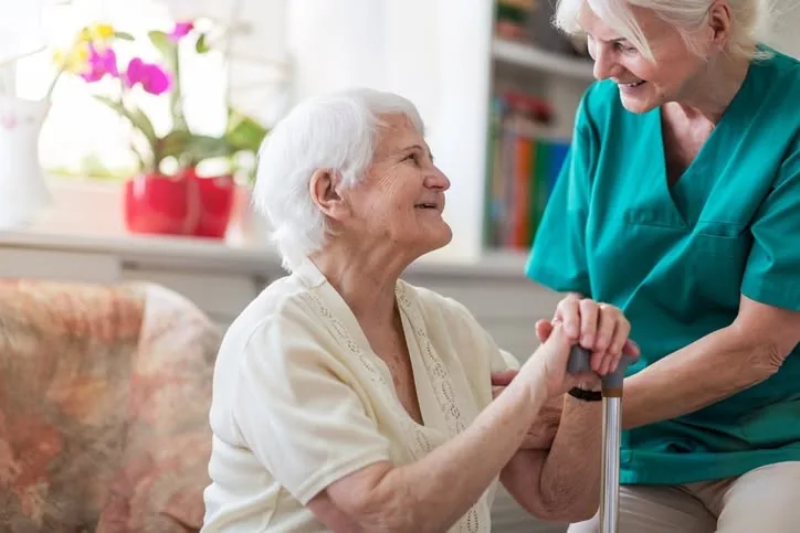 A caregiver in scrubs greeting an older woman with a cane