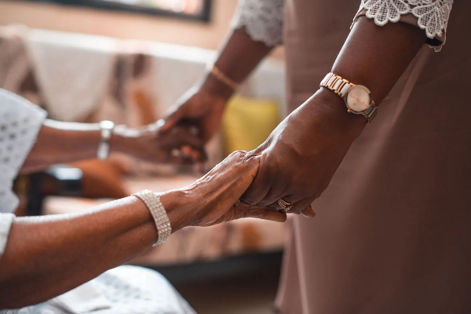 A younger woman holding an older woman's hands beside her armchair